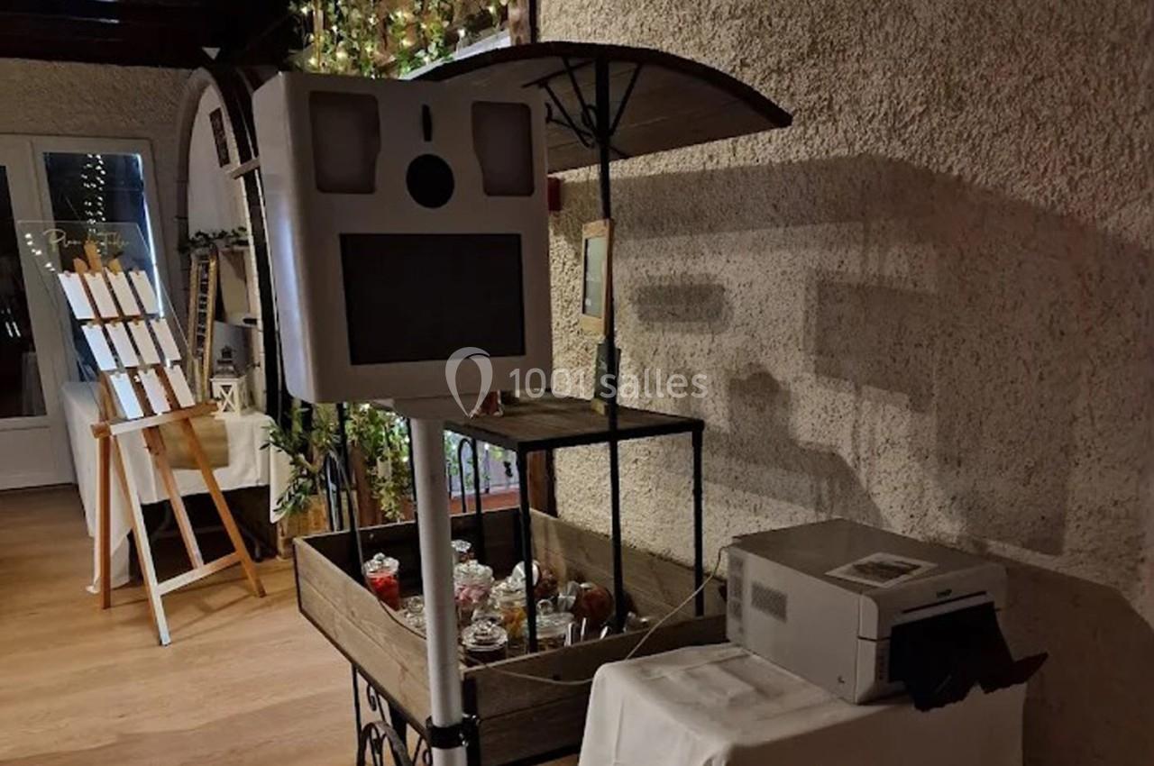 Borne photo avec écran et objectif, installée près d'une imprimante et d'un chariot décoratif dans un espace intérieur.