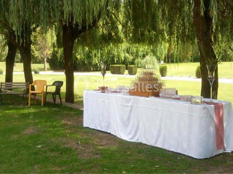 Table dressée avec une nappe blanche sous des arbres, contenant une pyramide de verres et des accessoires de réception.