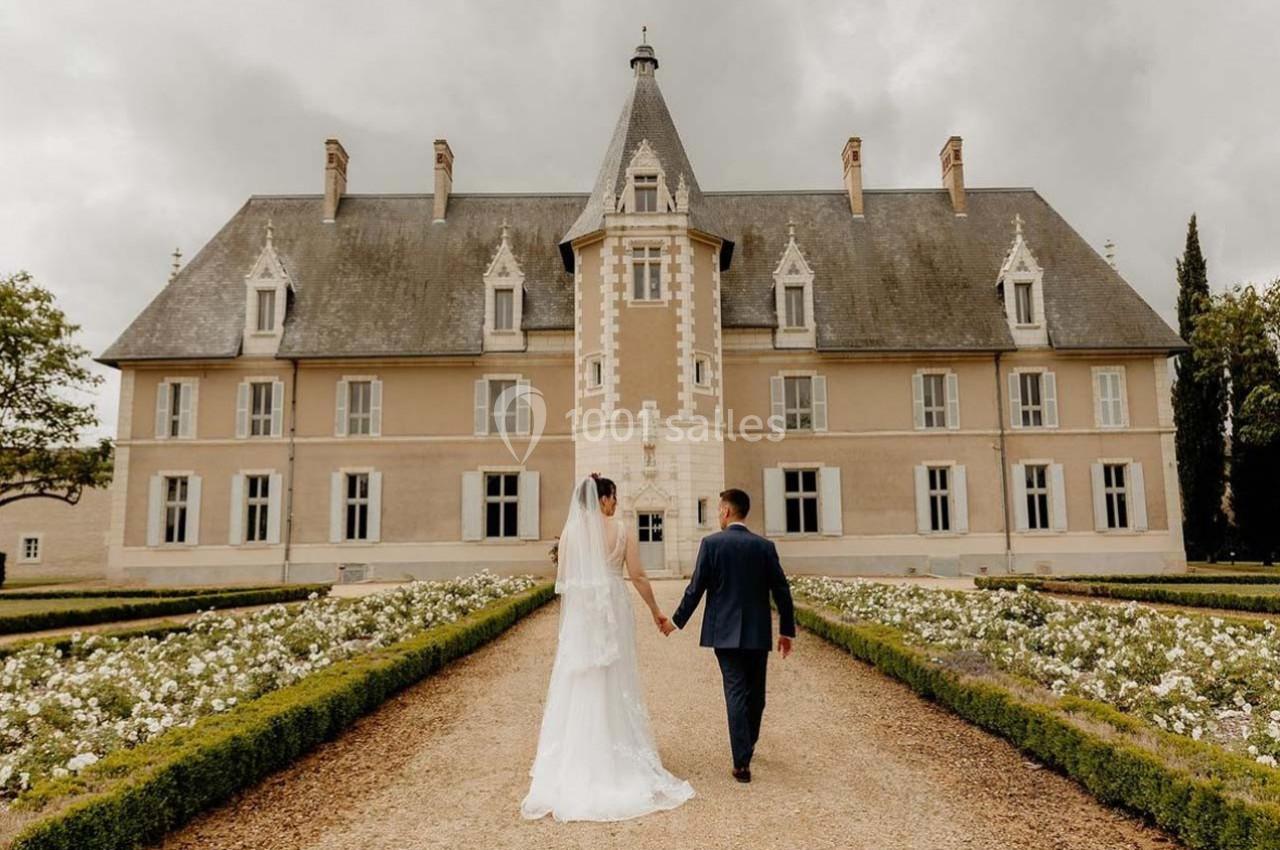 Un couple en tenue de mariage marche main dans la main devant un château entouré de jardins fleuris.