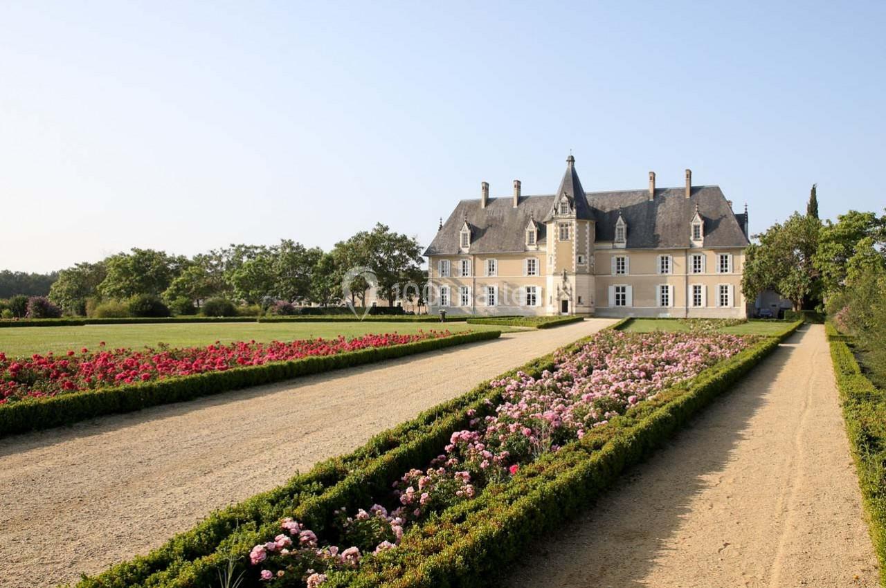 Allée bordée de parterres fleuris menant à un château en pierre entouré de jardins sous un ciel dégagé.