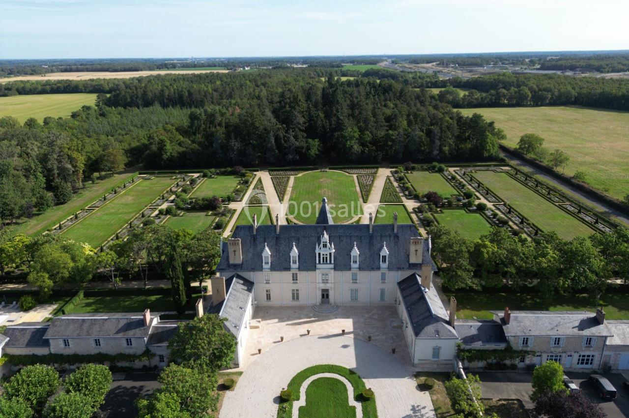 Vue aérienne d'un château entouré de jardins symétriques, de pelouses et de forêts sous un ciel dégagé.