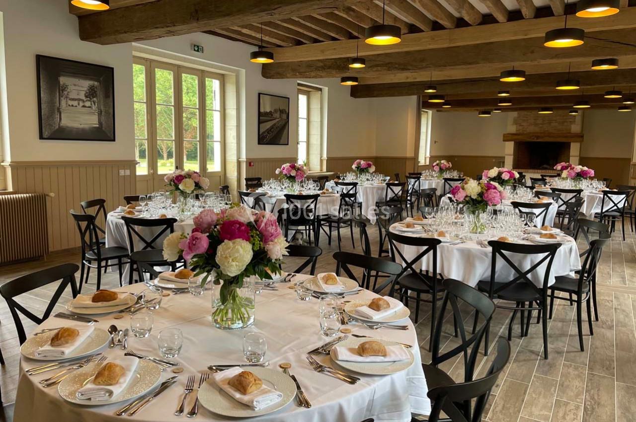 Salle de réception lumineuse avec tables rondes dressées, décorées de bouquets de fleurs et entourées de chaises noires.