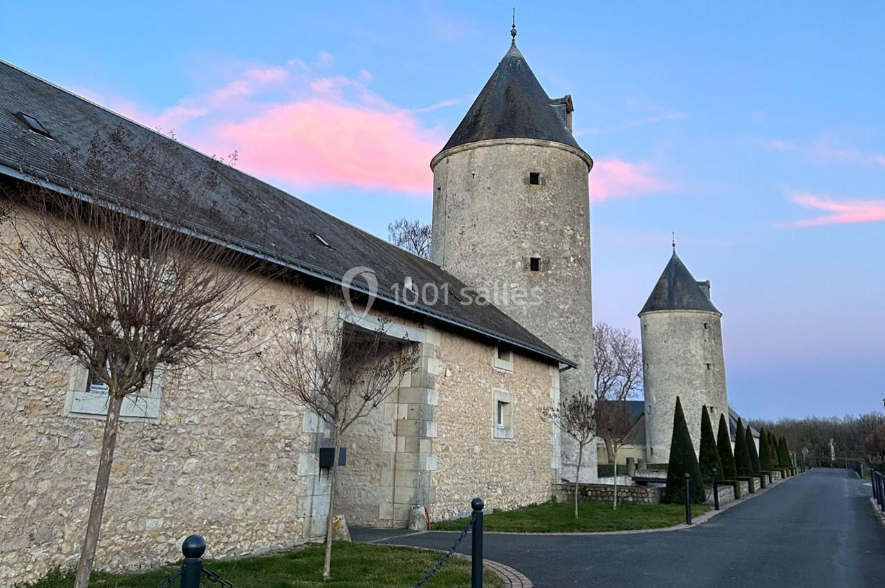 Deux tours en pierre et un bâtiment adjacent sous un ciel bleu avec des nuages roses, bordés d'arbres et d'une allée.