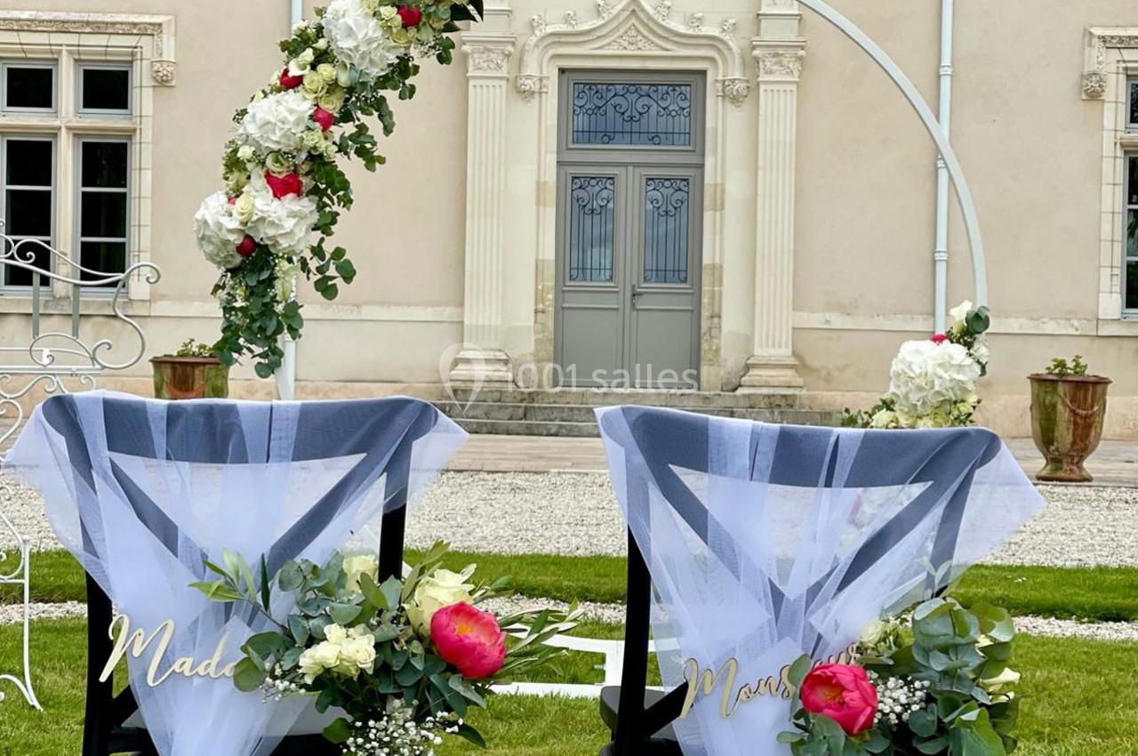 Deux chaises décorées de voiles blancs et de fleurs devant une arche florale, dans un jardin près d'un bâtiment ancien.