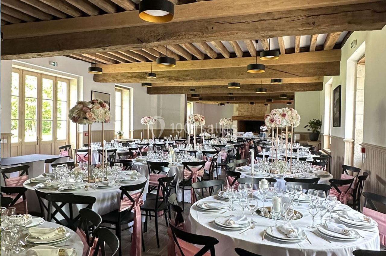 Salle de réception décorée pour un événement, avec des tables rondes dressées et des centres de table fleuris.