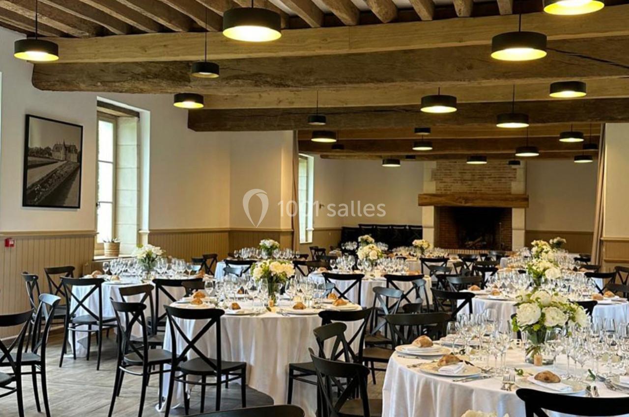 Salle de réception élégamment décorée avec des tables rondes dressées, des fleurs blanches et un plafond en poutres…
