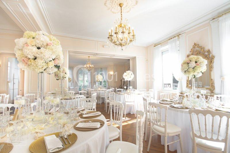 Salle de réception lumineuse décorée avec des tables rondes, des chaises blanches et des compositions florales élégantes.