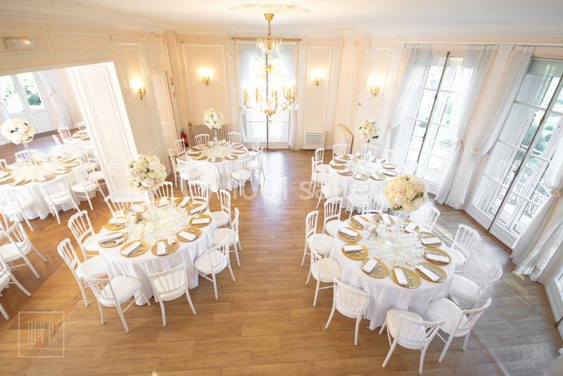 Salle lumineuse avec tables rondes dressées, nappes blanches, chaises blanches et décorations florales élégantes.