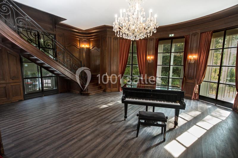 Salle élégante avec parquet, piano à queue noir, escalier en fer forgé, boiseries et lustre en cristal.