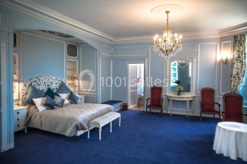 Chambre élégante avec murs bleus, lit double, lustre, fauteuils rouges et moquette bleue.