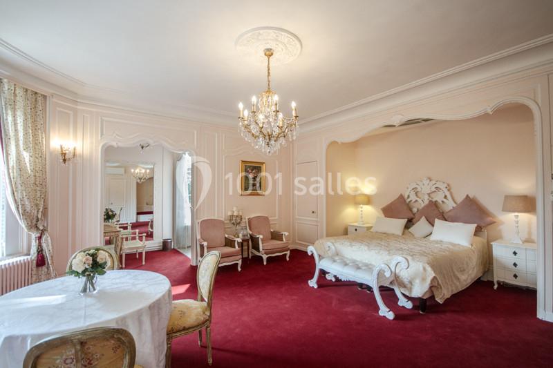 Chambre élégante avec lit double, lustre, tapis rouge, table ronde et fauteuils dans un décor classique.