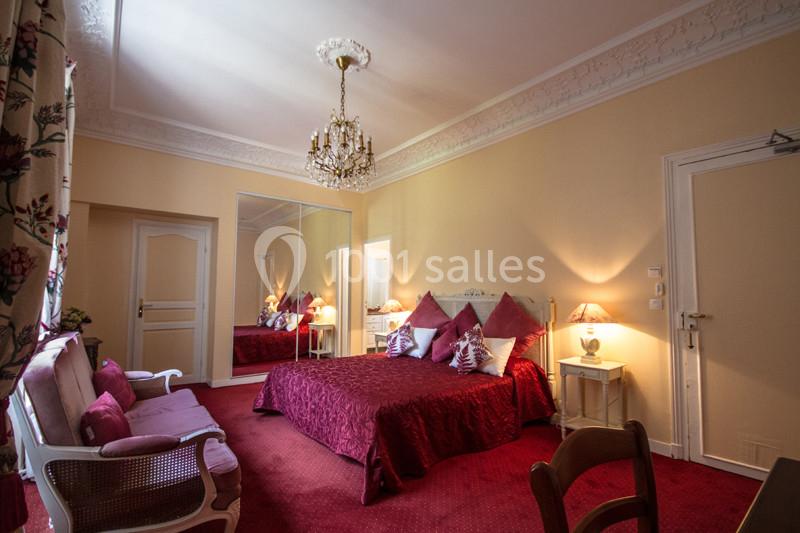 Chambre avec lit double, couvre-lit rouge, fauteuils assortis, lustre et miroir reflétant la pièce.