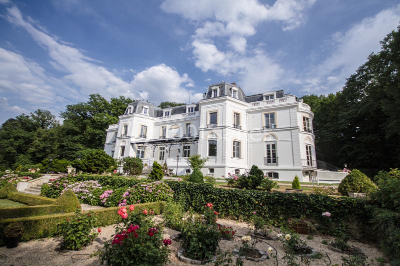 Façade d'un château blanc entouré de jardins fleuris et d'arbustes, sous un ciel partiellement nuageux.