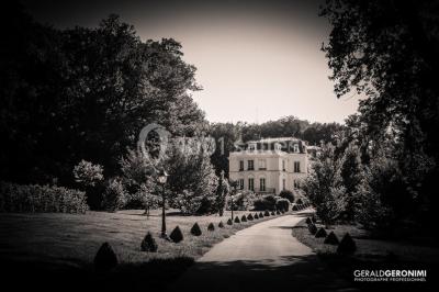 Allée bordée d'arbustes menant à une grande maison entourée d'arbres, capturée en noir et blanc.