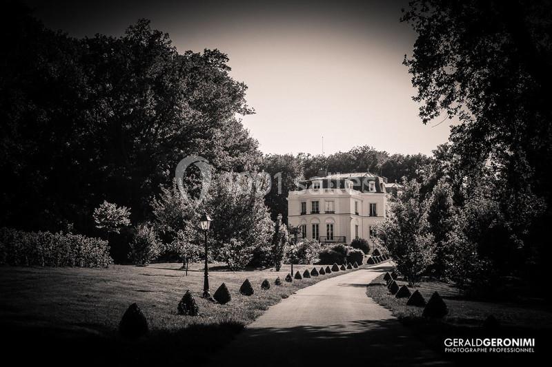 Allée bordée d'arbustes menant à une grande maison entourée d'arbres, capturée en noir et blanc.