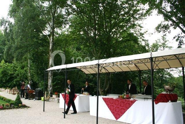 Des tables de réception avec nappes rouges et blanches sont disposées sous un auvent dans un jardin arboré.
