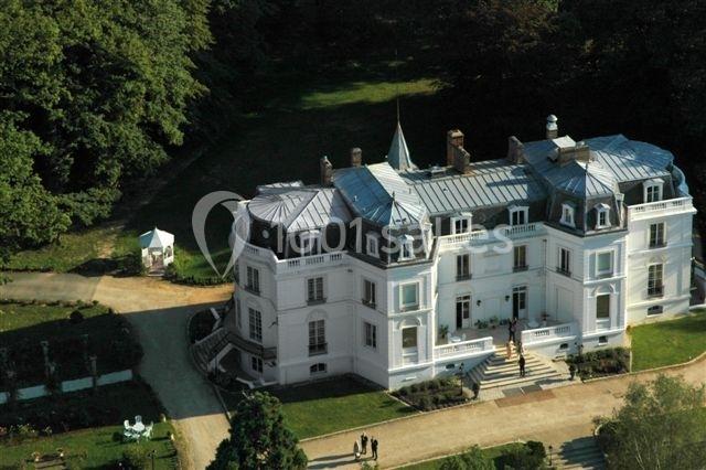 Vue aérienne d'un château blanc entouré de jardins et d'arbres, avec une allée menant à l'entrée principale.