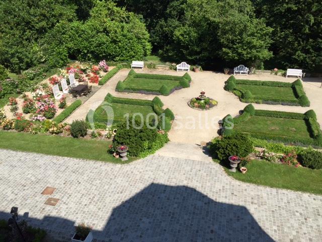 Vue aérienne d'un jardin symétrique avec parterres de fleurs, allées en gravier et bancs blancs entourés de verdure.