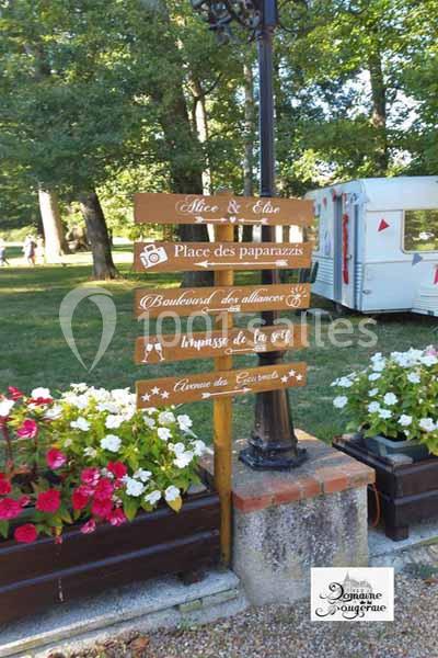 Panneau en bois indiquant différentes directions dans un parc, avec une caravane et des arbres en arrière-plan.