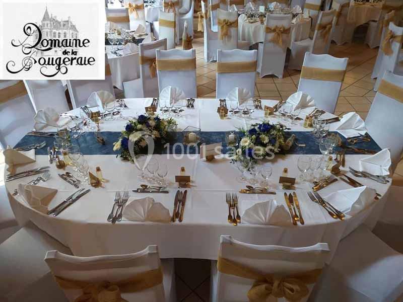 Salle de réception décorée avec des tables dressées, nappes blanches, chaises ornées de rubans dorés et centres de table…