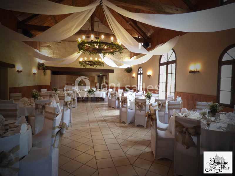 Salle de réception décorée avec des nappes blanches, des chaises ornées de rubans et un lustre central entouré de verdure.