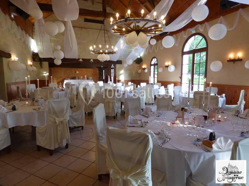 Salle de réception décorée pour un événement, avec tables rondes dressées, chaises couvertes et suspensions blanches.