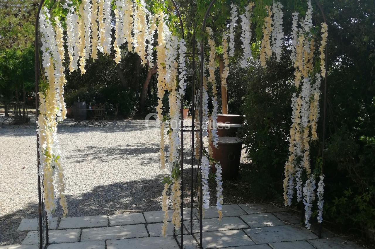 Arche métallique décorée de fleurs blanches suspendues, installée sur un sol pavé dans un jardin ensoleillé.
