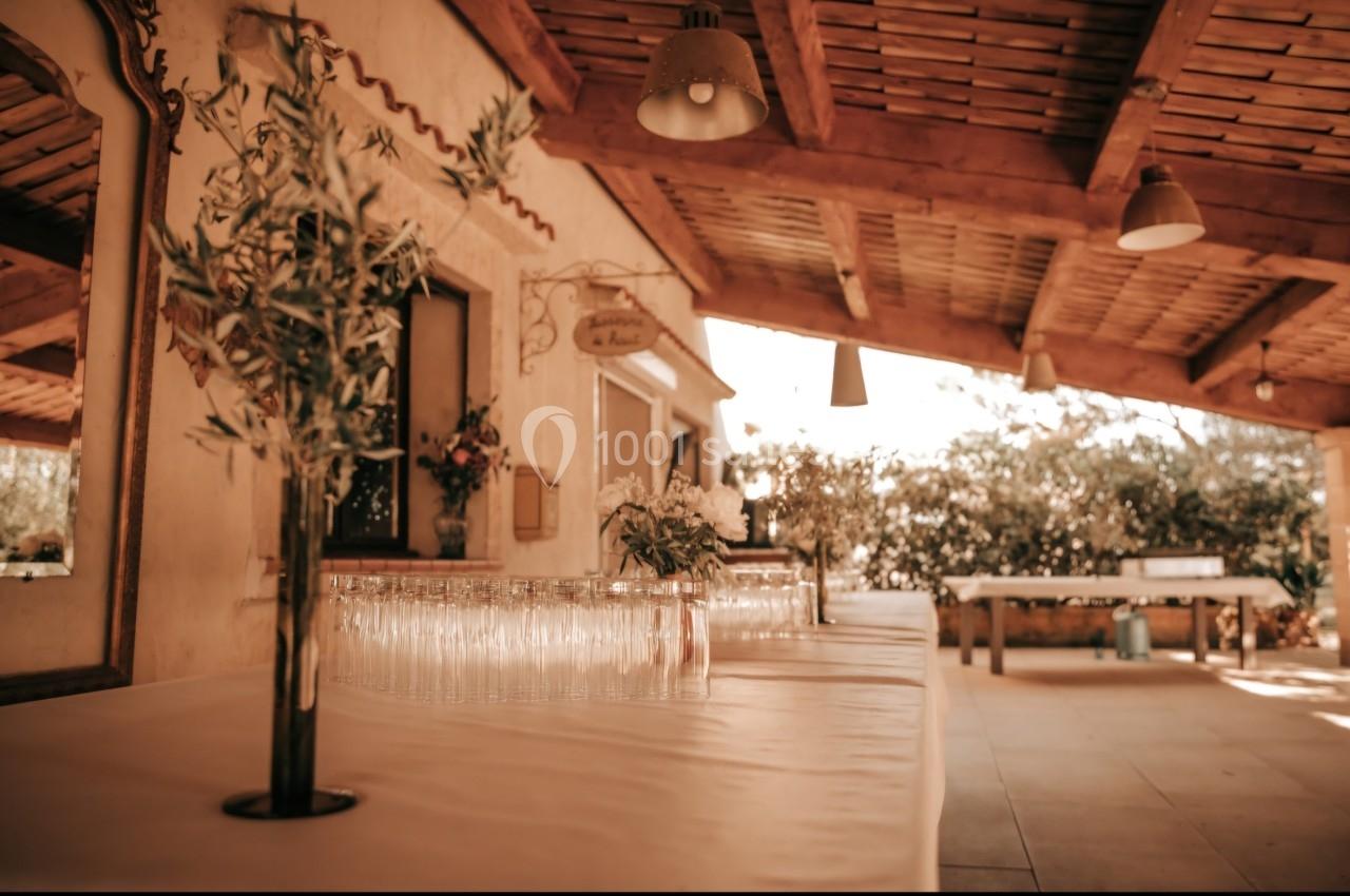 Table dressée avec des verres alignés sous une pergola en bois, décorée de plantes et éclairée par des suspensions.
