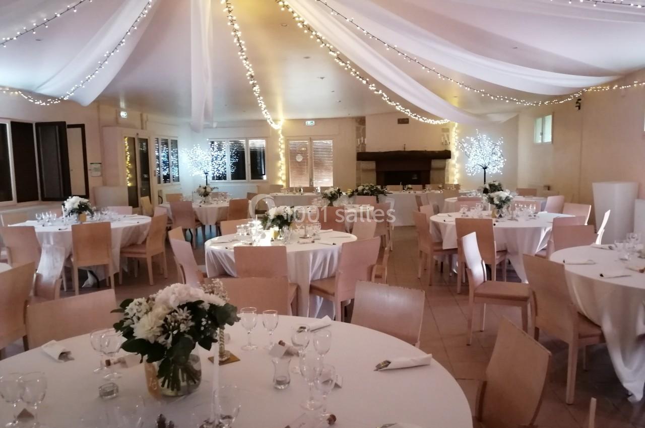 Salle de réception décorée avec des tables rondes, nappes blanches, fleurs et guirlandes lumineuses suspendues.