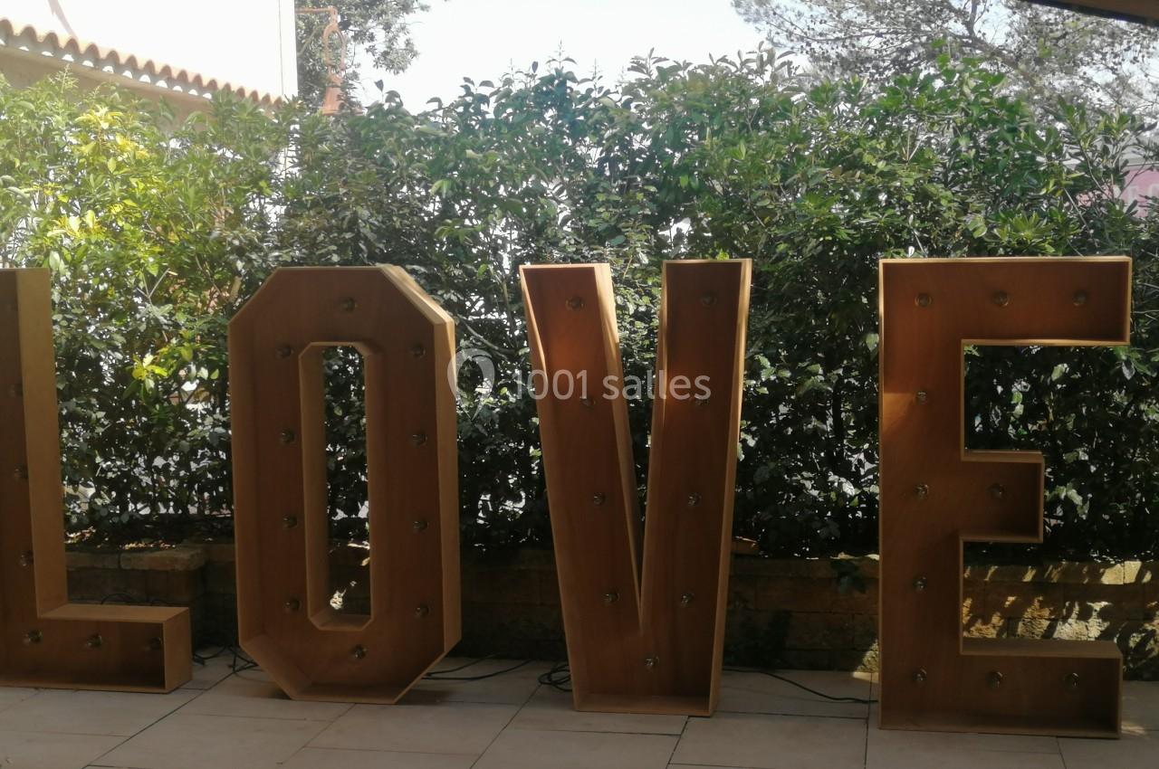 Lettres géantes en bois formant le mot ’LOVE’, éclairées par des ampoules, devant une haie verdoyante.