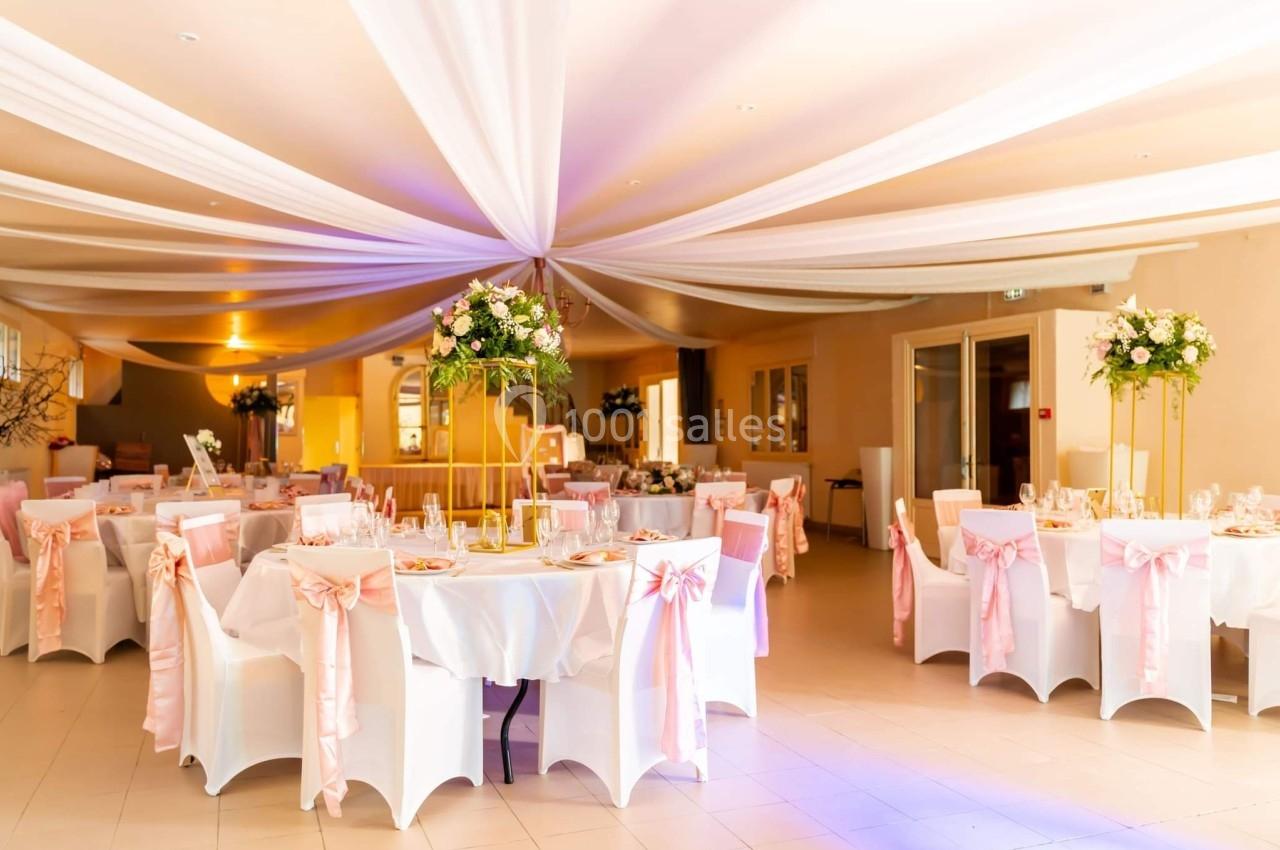 Salle de réception décorée avec des tables rondes, nappes blanches, chaises ornées de rubans roses et arrangements floraux.