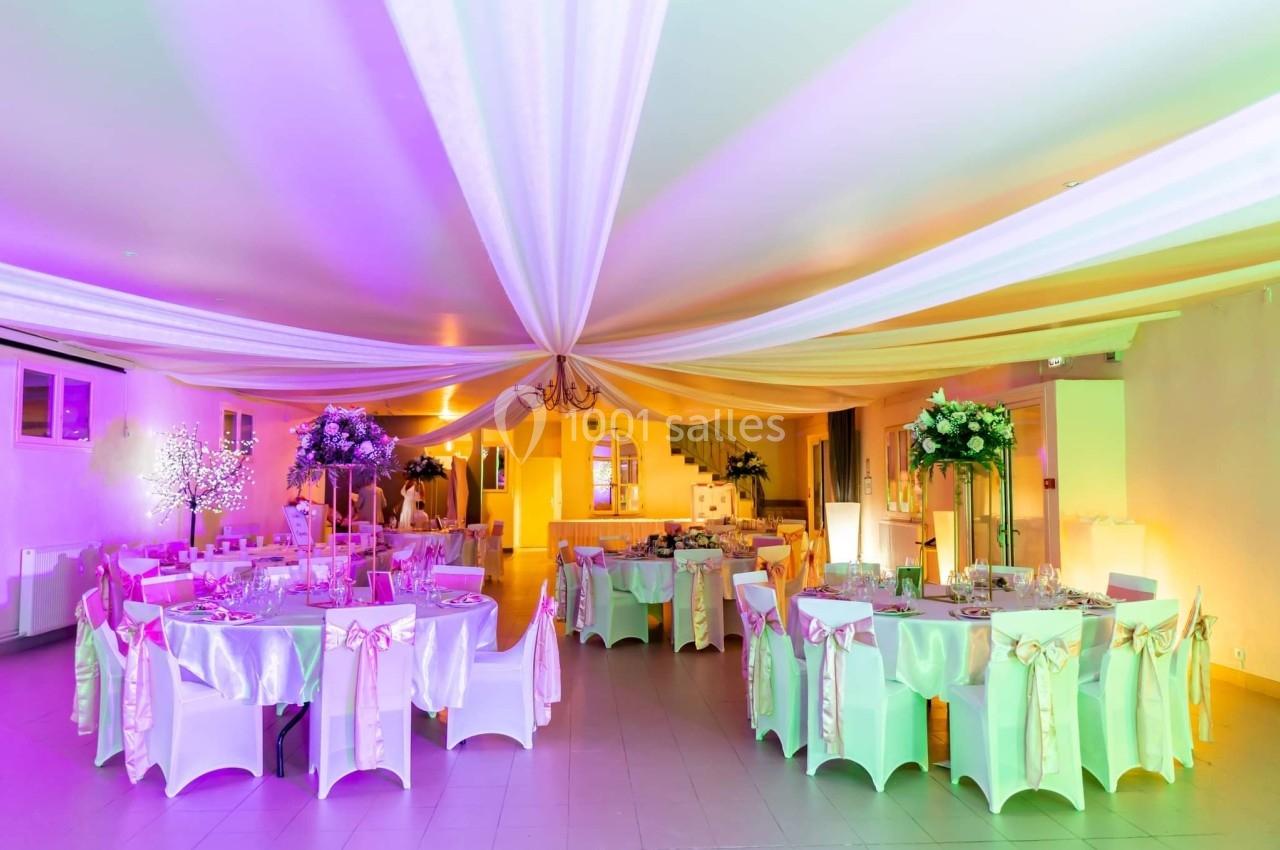 Salle de réception décorée avec des tables rondes, nappes blanches, rubans roses et éclairage coloré.