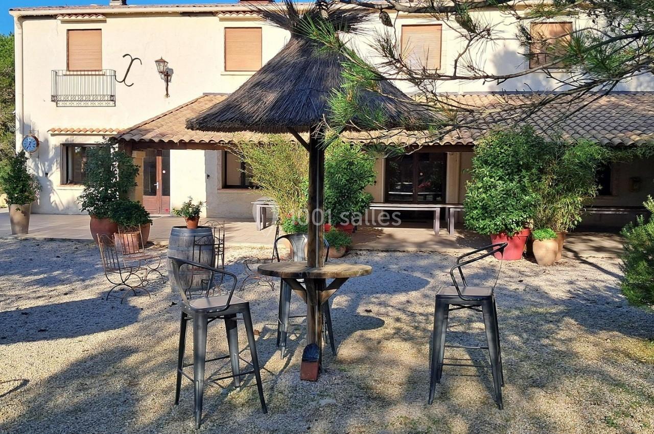 Terrasse ensoleillée avec table en bois, chaises hautes et parasol en chaume devant une maison à volets marron.