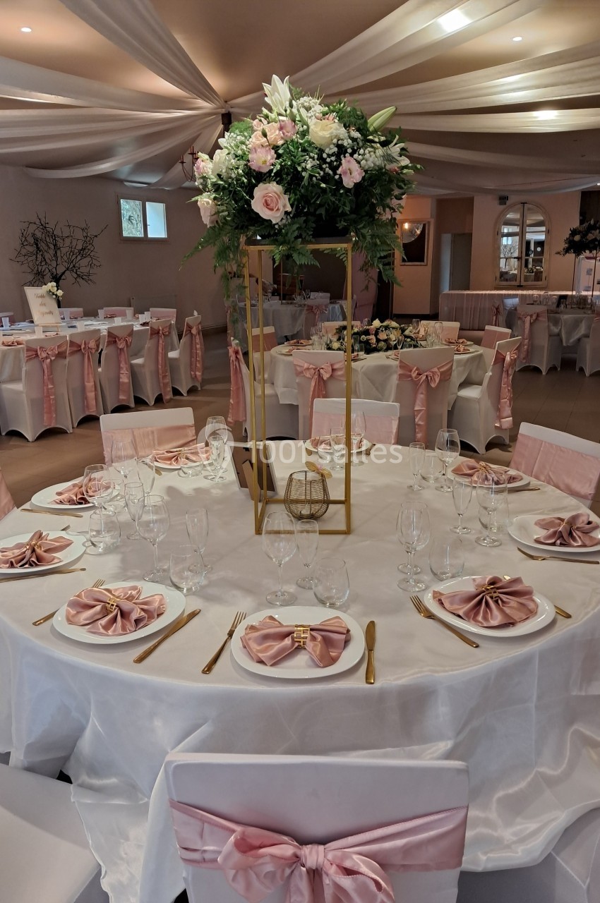 Salle de réception décorée avec des tables rondes, nappes blanches, serviettes roses pliées et centre de table floral.
