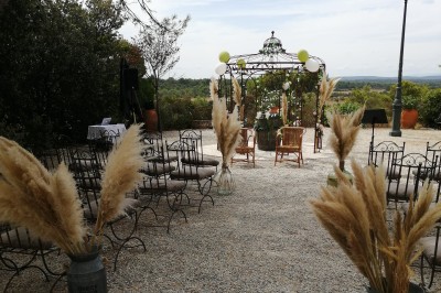 Cour extérieure ensoleillée avec des plantes en pots, des chaises en métal et un bâtiment à toit de tuiles en arrière-plan.