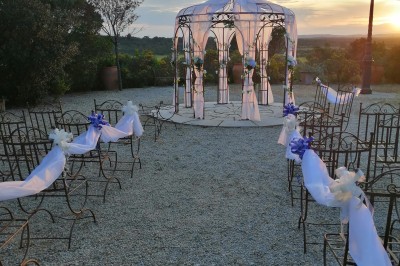 Cour extérieure ensoleillée avec des plantes en pots, des chaises en métal et un bâtiment à toit de tuiles en arrière-plan.