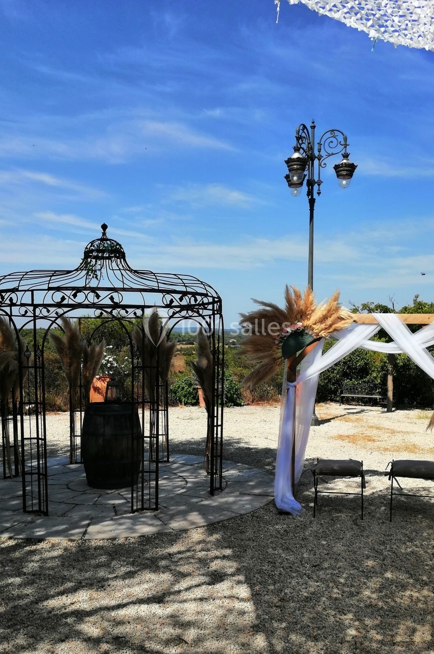 Structure métallique décorative et arche ornée de voilages et de fleurs séchées dans un espace extérieur ensoleillé.