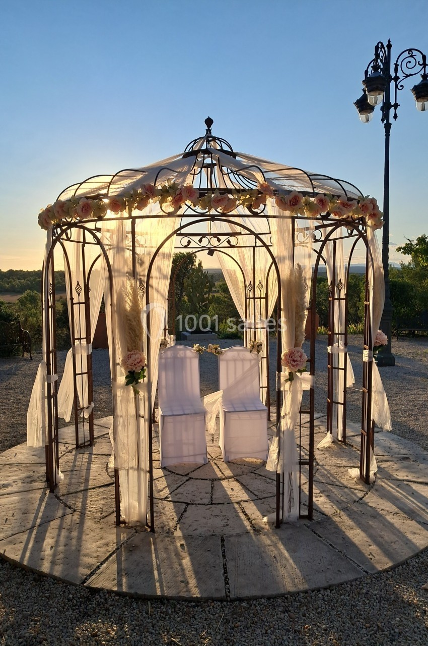 Tonnelle en fer forgé décorée de voilages et de fleurs, éclairée par le soleil couchant dans un jardin.