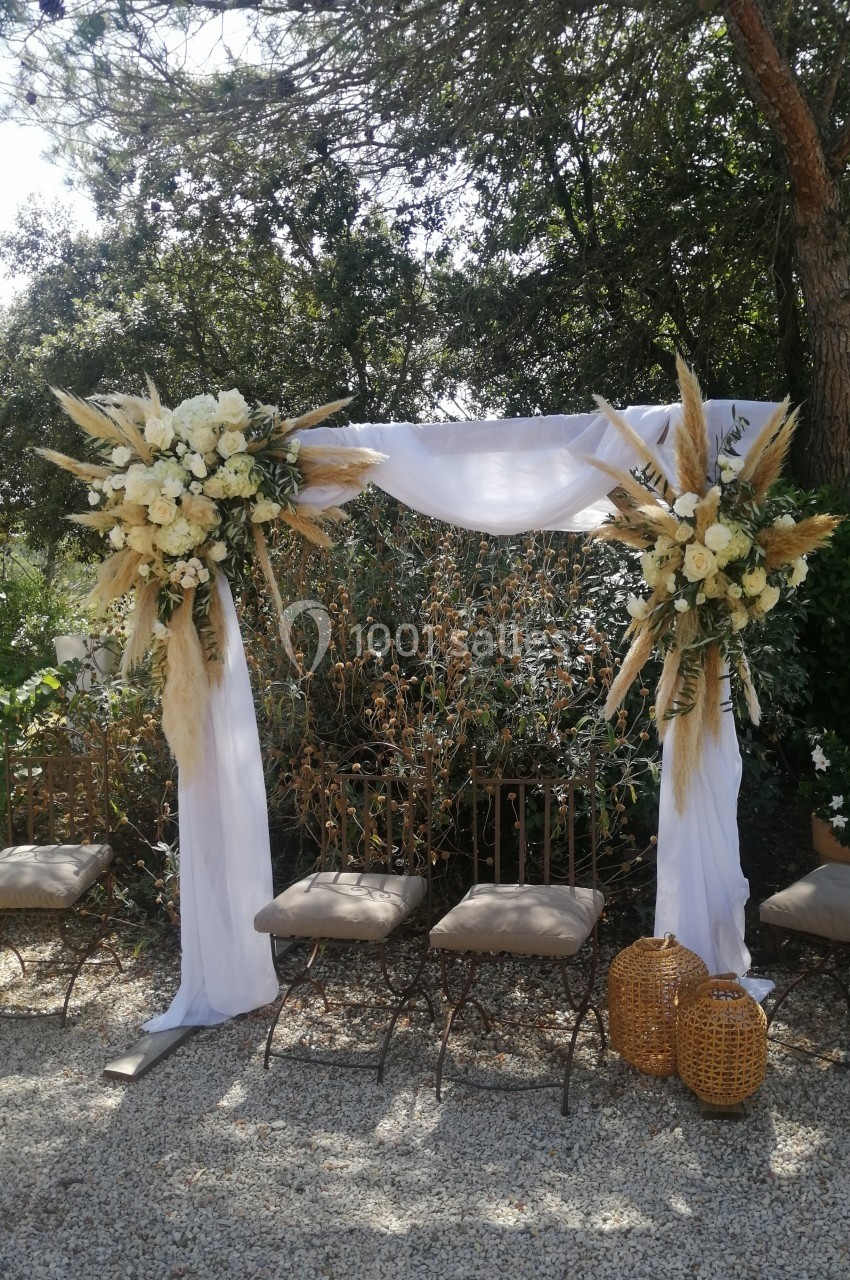 Arche de mariage décorée de fleurs blanches et de pampas, entourée de chaises et de verdure en extérieur.
