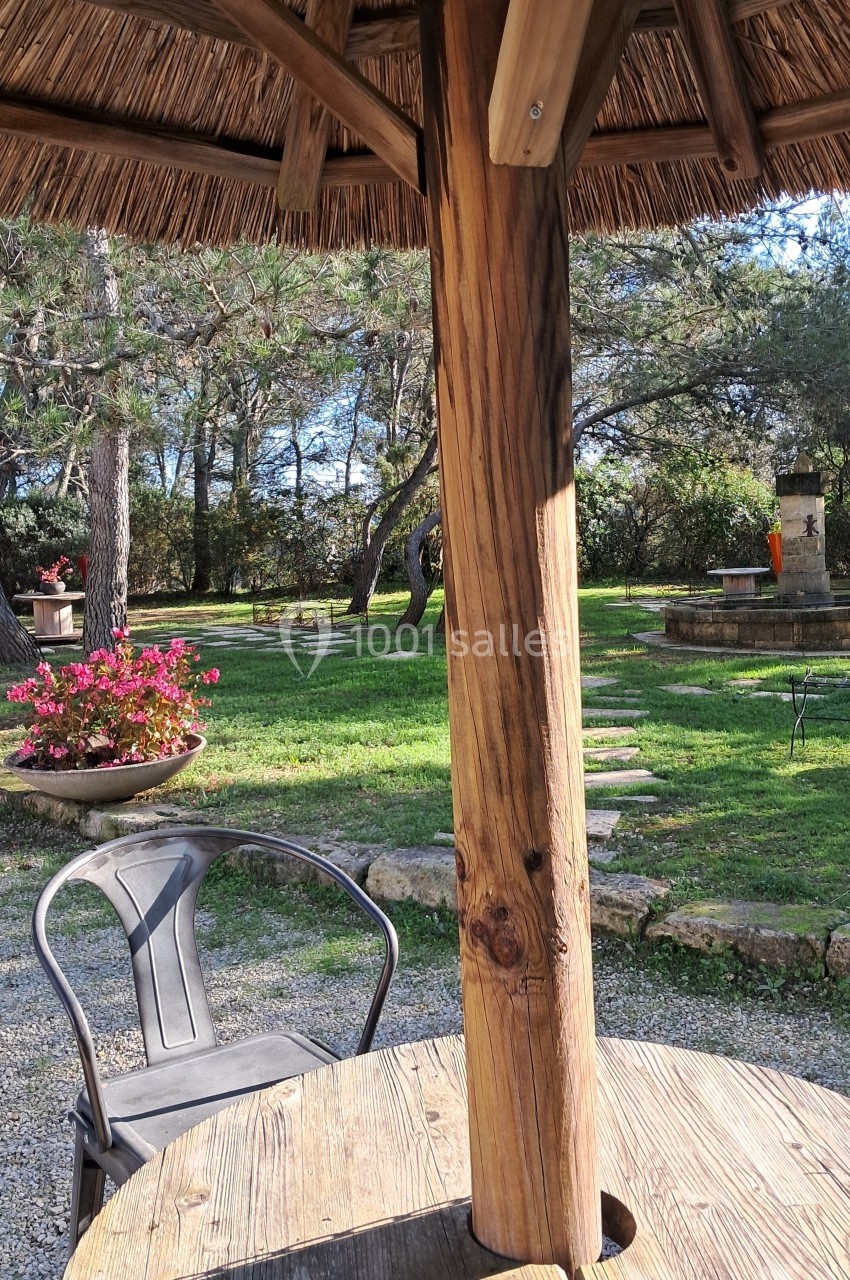Table en bois sous un parasol en chaume, chaise métallique et jardin arboré avec fleurs et fontaine en arrière-plan.