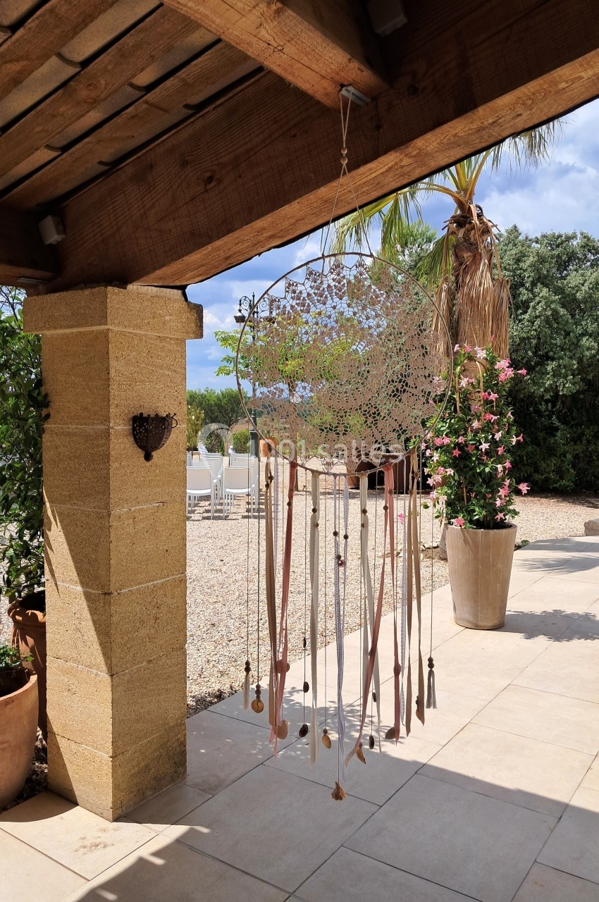 Attrait alt : Grand attrape-rêves suspendu sous une pergola en bois, entouré de plantes et d'une terrasse ensoleillée.