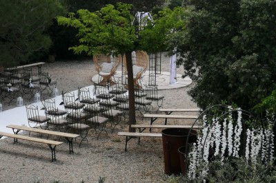 Cour extérieure ensoleillée avec des plantes en pots, des chaises en métal et un bâtiment à toit de tuiles en arrière-plan.