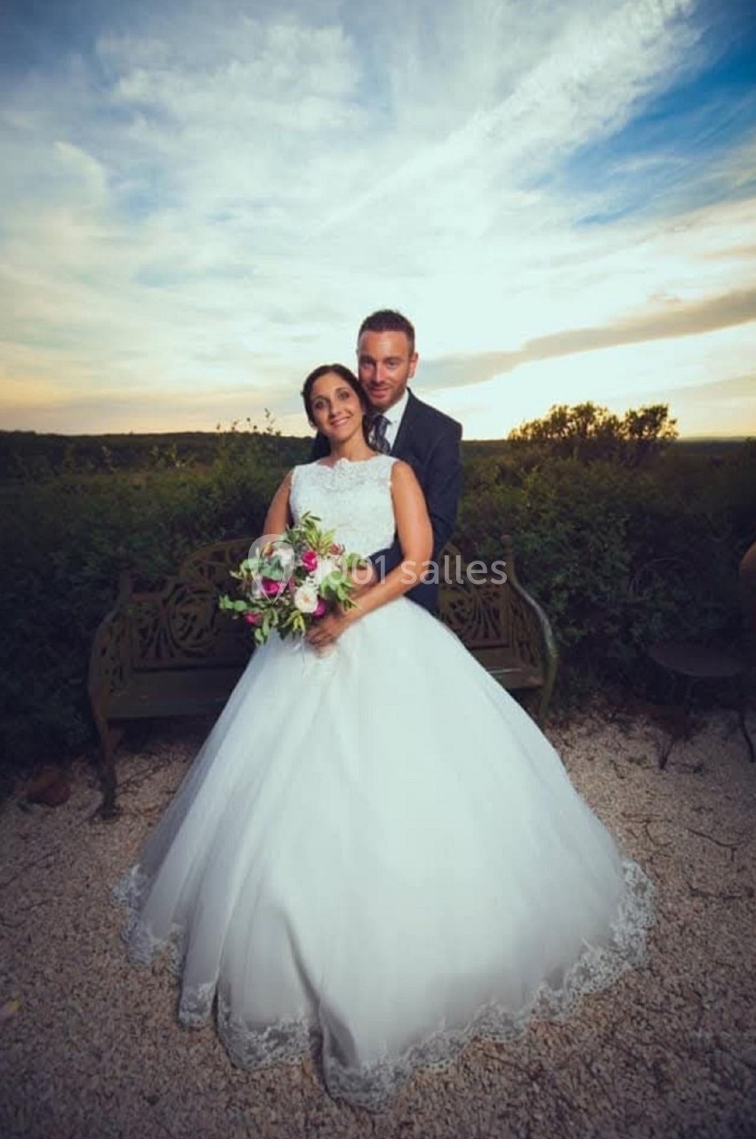 Un couple en tenue de mariage pose devant un banc dans un jardin au coucher du soleil.