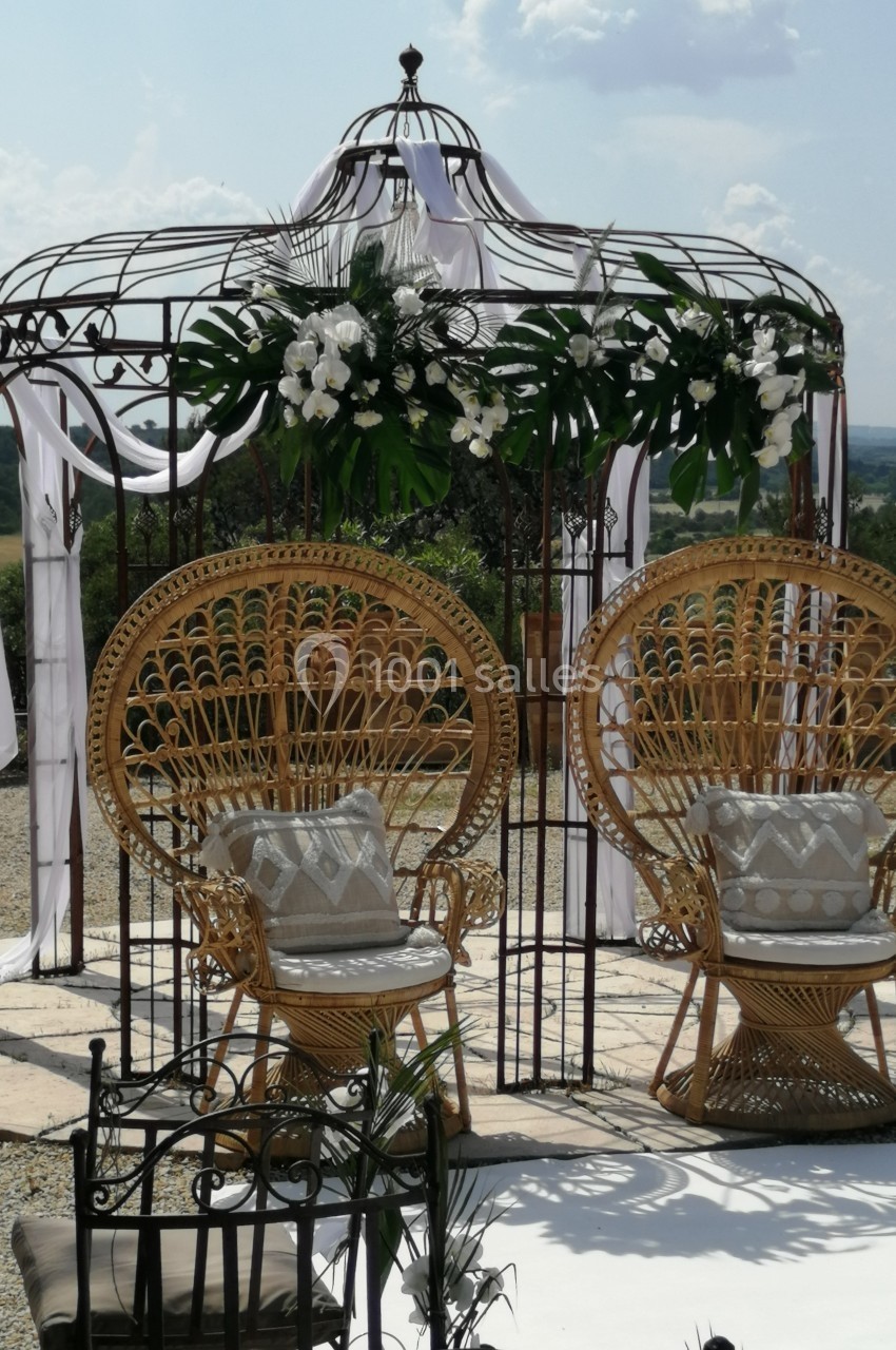 Deux fauteuils en rotin devant une arche métallique décorée de fleurs blanches et de voilages, en extérieur.