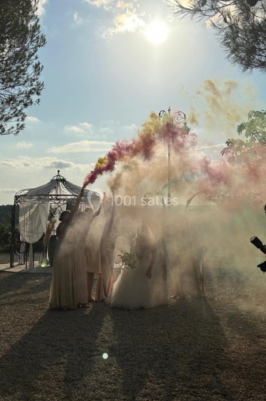 Des personnes en tenue claire lancent des fumigènes colorés dans un cadre extérieur ensoleillé près d'une tonnelle.