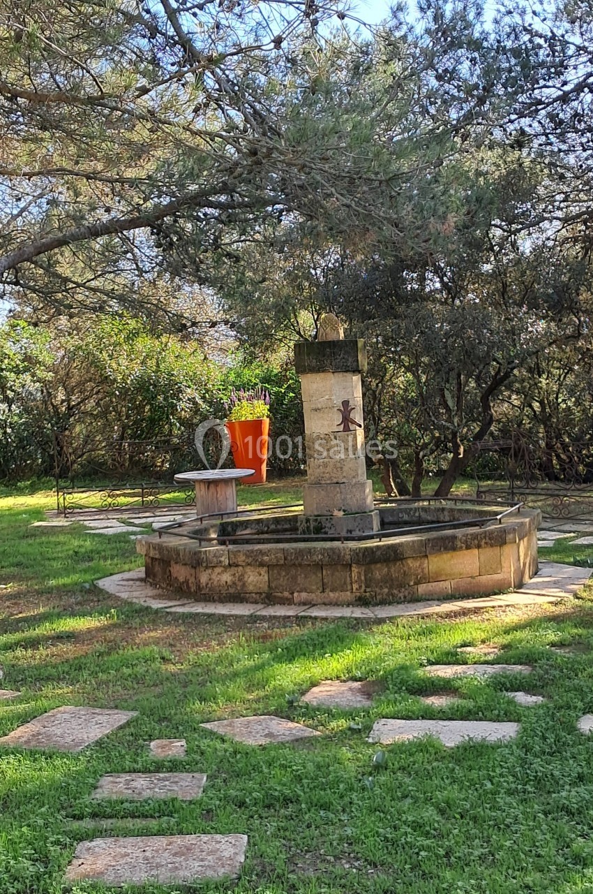 Fontaine en pierre entourée d'herbe et d'arbres, avec un grand pot orange en arrière-plan.
