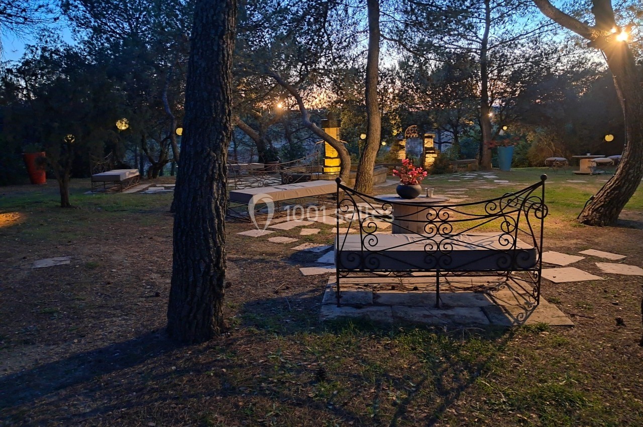 Banc en fer forgé dans un jardin boisé éclairé par des lampes suspendues au crépuscule.