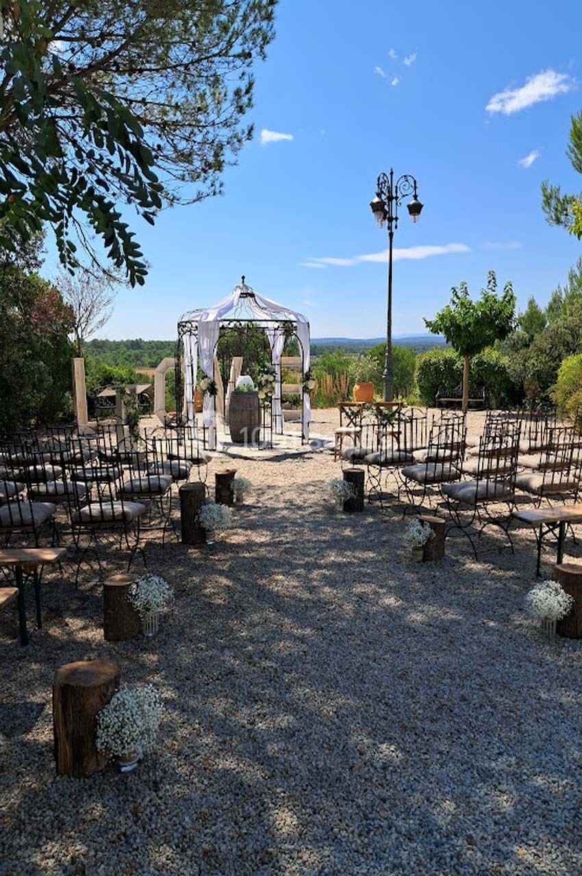 Chaises disposées en extérieur devant une arche décorée pour une cérémonie, entourées de végétation sous un ciel dégagé.