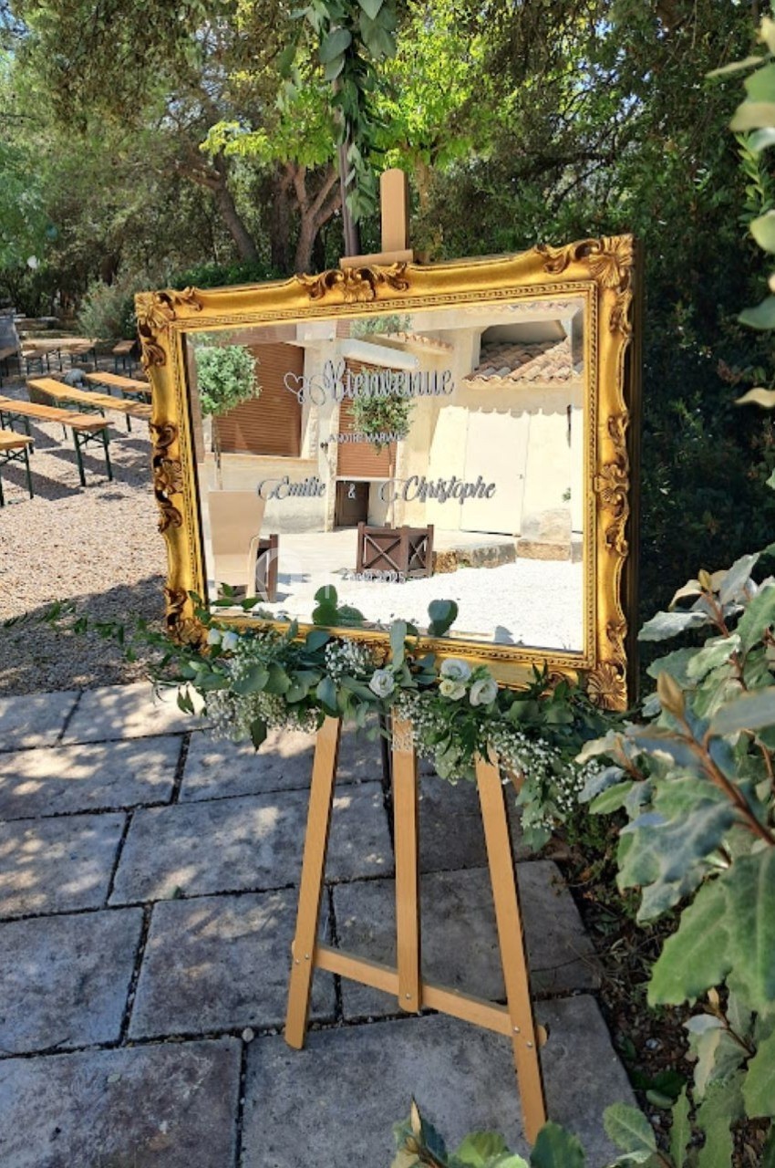 Miroir doré sur chevalet décoré de feuillage, reflétant une cour extérieure avec tables et végétation.