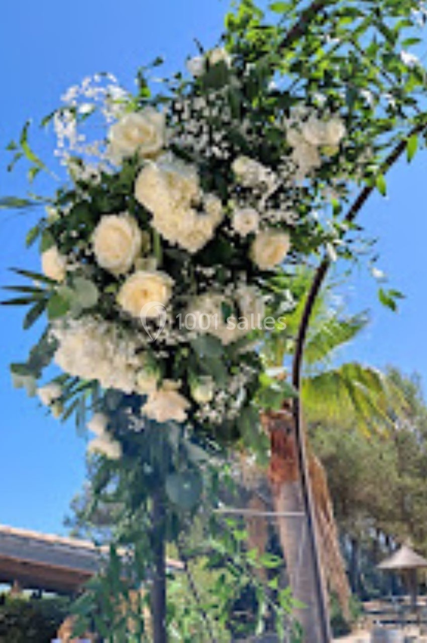 Arrangement floral blanc et vert suspendu sur une arche métallique, avec un ciel bleu et des arbres en arrière-plan.